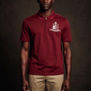 May include: Burgundy polo shirt with a silver crest design on the chest. The shirt has a zipper closure and short sleeves. The model is wearing khaki pants.