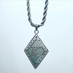 May include: A silver chain necklace with a diamond-shaped pendant featuring the letter "K" in a raised font. The pendant is accented with small red gemstones.