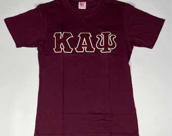 Op de afbeelding: Een maroon t-shirt met een grote witte omtrek van de letters "ΚΑΨ" in bordeaux.