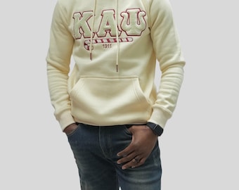 Puede incluir: Una sudadera con capucha de color crema con un diseño de bastón de caramelo rojo y blanco y el texto "KAU 1911" bordado en la parte delantera.