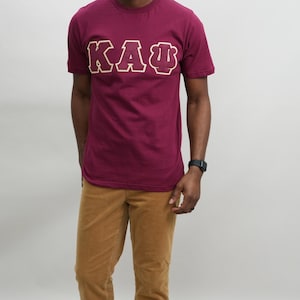 以下が含まれることがあります： 金色の輪郭で白い文字「KAΨ」が書かれた、マルーンのTシャツです。
