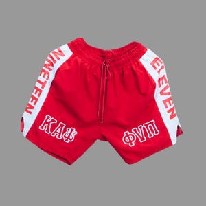 May include: Red athletic shorts with white side stripes. The shorts feature the words "NINETEEN" and "ELEVEN" in white on the sides, along with Greek letters. The shorts have a drawstring waist.