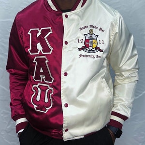 May include: A white and maroon satin varsity jacket with a Kappa Alpha Psi fraternity crest and the letters "KAU" on the left side. The crest features a shield with a compass, a key, and a torch. The text "Kappa Alpha Psi 1911 Fraternity, Inc." is embroidered on the jacket.