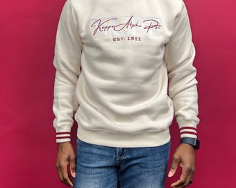 Puede incluir: Una sudadera de color crema con cuello redondo y rayas marrones en los puños. La sudadera tiene un diseño bordado en marrón que dice "Kappa Alpha Psi EST. 1911".