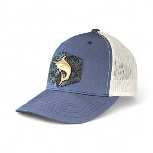 May include: A blue baseball cap with a white mesh back. The cap features a decorative patch with a marlin design in gold and a floral pattern. The brim is curved, and the cap has a button on top.