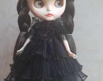 May include: A black doll with long black hair styled in braids, wearing a black lace dress with multiple layers of ruffles. The doll has green eyes and is wearing black shoes.
