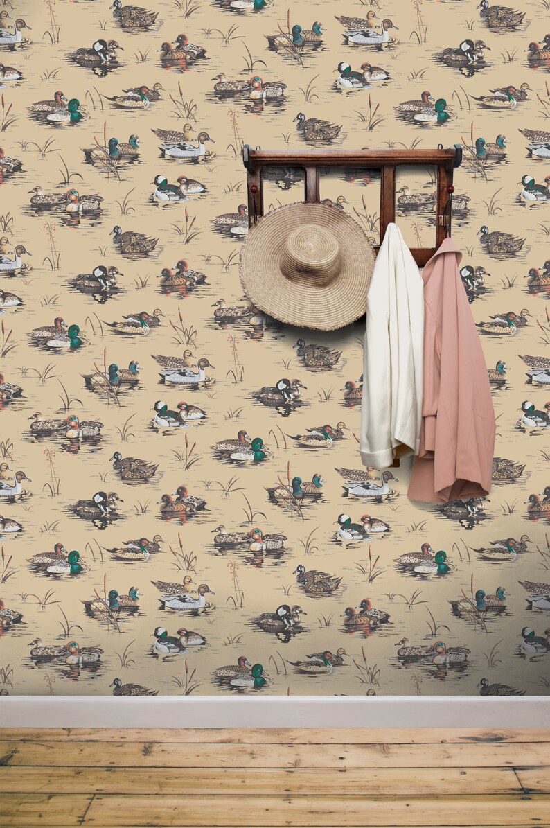 May include: A beige wallpaper with a repeating pattern of ducks swimming in a pond. A wooden coat rack with a straw hat and two jackets hanging on it is in the center of the image.