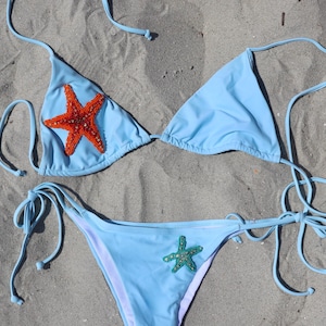 Può includere: Bikini azzurro con decorazioni a stella marina. Il top a triangolo presenta una grande stella marina arancione, mentre la parte inferiore ha una stella marina verde più piccola. Il bikini è realizzato in tessuto liscio e ha chiusure con lacci.