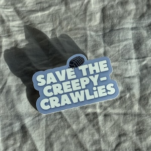 May include: A light blue sticker with the text "SAVE THE CREEPY-CRAWLIES" in white, featuring a small illustration of a pill bug. The sticker is on a wrinkled, light gray fabric background.