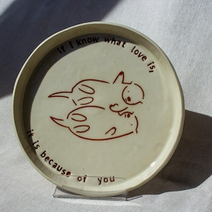 May include: A round, off-white ceramic plate with a brown illustration of two stylized cats. The plate has the inscription "If I know what love is, it is because of you" in brown lettering. The plate is displayed on a clear stand.