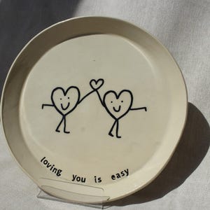 Op de afbeelding: Crèmekleurige keramische plaat met een zwarte, handgetekende illustratie van twee hartvormige figuren die elkaars handen vasthouden. Op de plaat staat de zin "loving you is easy" in het zwart. Een charmant, grillig ontwerp.