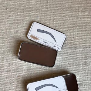 May include: Two small metal tins with lids containing eyebrow wax. The tins are labeled "Light .25 oz" and "Brown .25 oz". Each tin has a small illustration of an eyebrow shape.