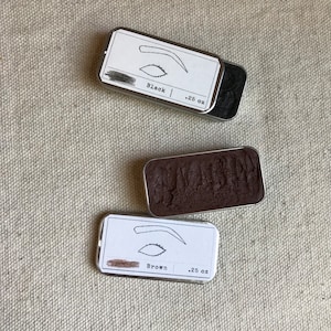 Puede incluir: Dos pequeñas latas de metal con tapas que contienen maquillaje para cejas. La lata de la izquierda está etiquetada como "Black .25 oz" y la lata de la derecha está etiquetada como "Brown .25 oz".