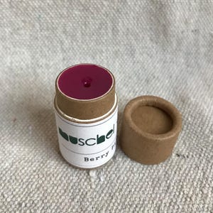 Puede incluir: Un bálsamo labial de color bayas en un envase cilíndrico de cartón. El envase tiene una etiqueta blanca con el nombre de la marca y "Berry" impreso. La tapa está al lado del envase abierto. El bálsamo es visible.