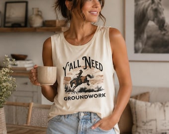 May include: A cream-colored sleeveless tank top featuring a graphic print of a person riding a horse in a desert landscape with cacti. The text Y'ALL NEED GROUNDWORK is printed in black lettering above and below the central illustration.