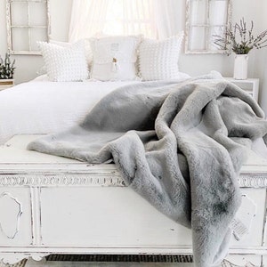 以下が含まれることがあります： 白いベッドに、グレーのフェイクファーの毛布がかけられています。毛布はベッドの端から床まで垂れ下がっています。ベッドは白いシーツと枕でできています。ベッドの足元には白いチェストがあります。