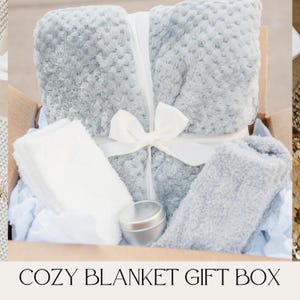 May include: A gift box filled with cozy items, including a gray plush blanket, a white fluffy towel, a gray fuzzy slipper sock, and a small silver tin.