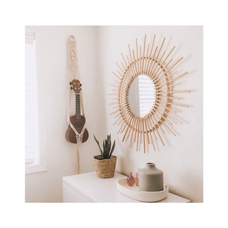 May include: A white wall with a wooden sunburst mirror, a ukulele hanging from a macrame wall hanging, a potted plant, and a white dresser with a white tray and glass bottles on top.