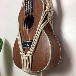 May include: A brown ukulele with a white trim is hanging on a wall using a macrame wall hanger made of natural colored cotton rope.