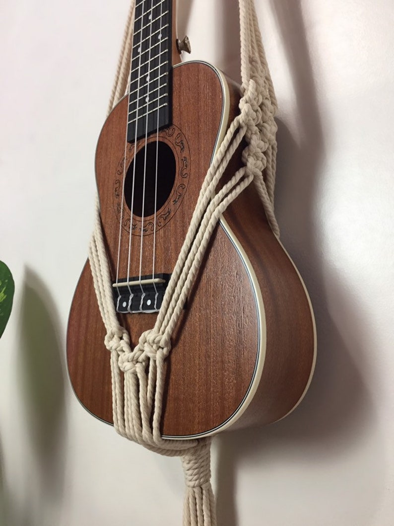 May include: A brown ukulele with a white trim is hanging on a wall using a macrame wall hanger made of natural colored cotton rope.