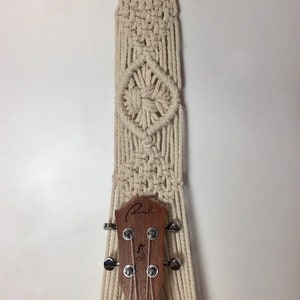 May include: A white macrame wall hanging with a wooden ring at the top. The hanging is designed to hold a ukulele, which is partially visible in the image. The ukulele has a wooden body and a black fretboard.