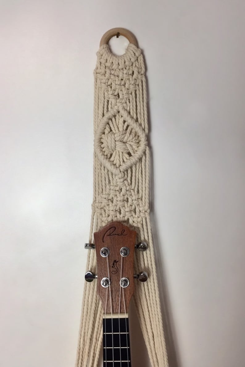 May include: A white macrame wall hanging with a wooden ring at the top. The hanging is designed to hold a ukulele, which is partially visible in the image. The ukulele has a wooden body and a black fretboard.