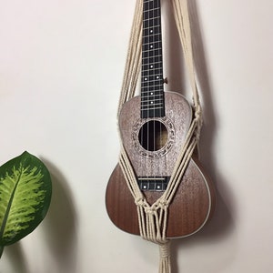 May include: A white macrame wall hanging with a brown ukulele hanging from it. The ukulele is hanging on a white wall.