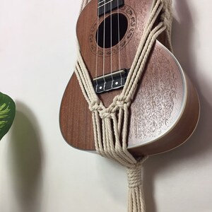 May include: A brown ukulele with a white rope macrame wall hanging. The ukulele has a decorative design around the sound hole.