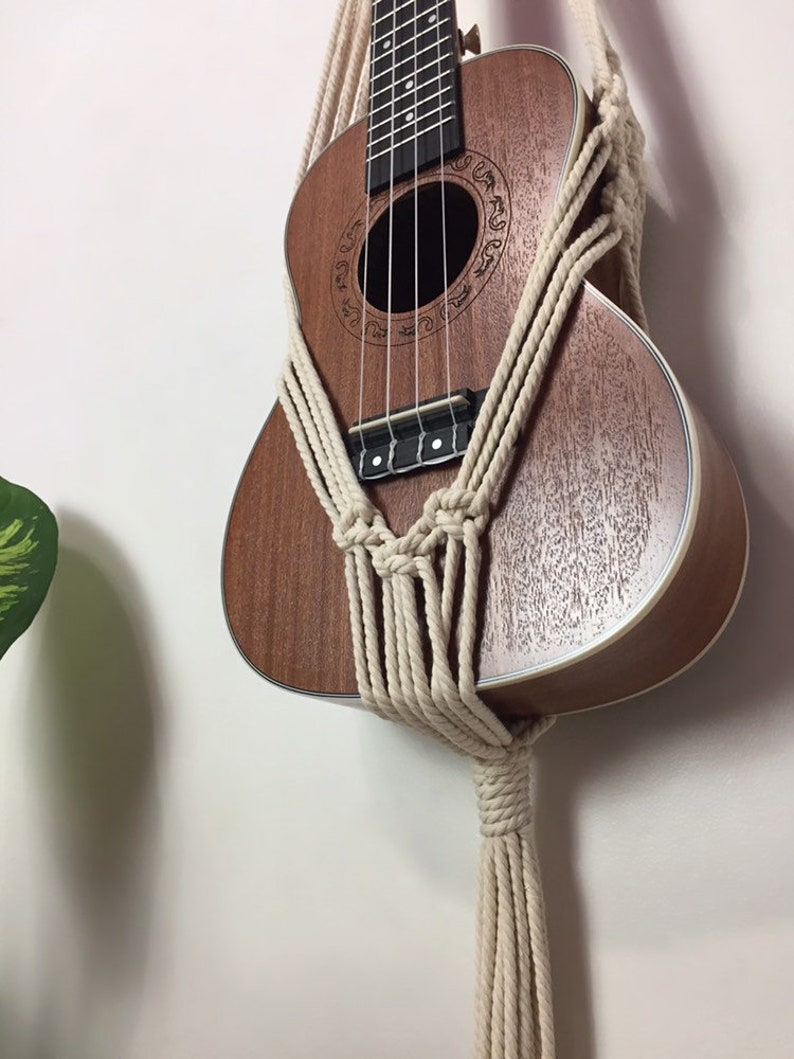 May include: A brown ukulele with a white rope macrame wall hanging. The ukulele has a decorative design around the sound hole.
