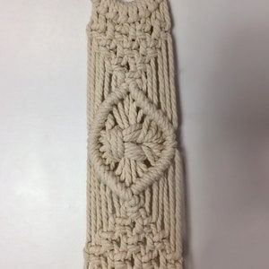 May include: A macrame plant hanger with a wooden ring at the top and a wooden sign at the bottom that says "Rock". The hanger is made of natural colored cotton cord and has a diamond pattern.