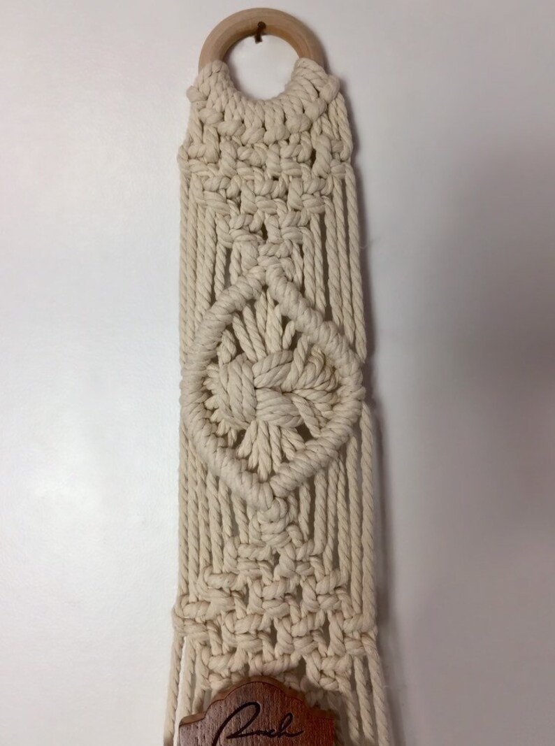 May include: A macrame plant hanger with a wooden ring at the top and a wooden sign at the bottom that says "Rock". The hanger is made of natural colored cotton cord and has a diamond pattern.