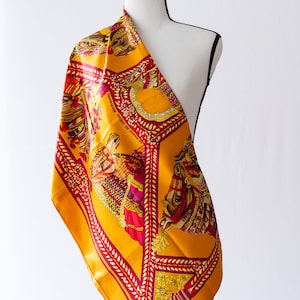 Pode incluir: Lenço de seda amarelo mostarda com um design equestre detalhado. O lenço apresenta um padrão repetido de cavalos, selas ornamentadas e o texto "LA DANSE DU CHEVAL MARWARI" em uma borda bordô. O lenço está drapeado sobre um manequim.