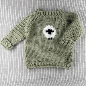 May include: A sage green knitted sweater with a white and black sheep design on the front. The sweater has long sleeves with ribbed cuffs and a ribbed hem. The neckline is also ribbed.