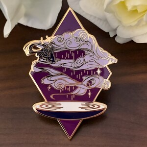 Pu&ograve; includere: Spilla in smalto viola e oro con un design a nuvola vorticosa e un cerchio bianco in basso. La spilla ha la forma di un diamante.