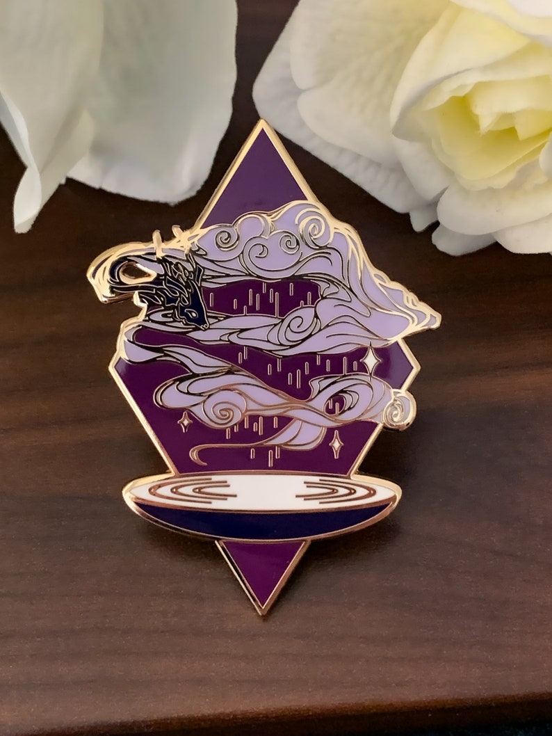 Pu&ograve; includere: Spilla in smalto viola e oro con un design a nuvola vorticosa e un cerchio bianco in basso. La spilla ha la forma di un diamante.