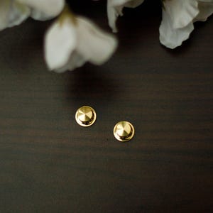 Pode incluir: Dois pequenos brincos de botão redondos dourados com um design canelado sobre uma superfície castanha escura. Pétalas de flores brancas são visíveis no fundo, adicionando um contraste suave aos brincos metálicos.