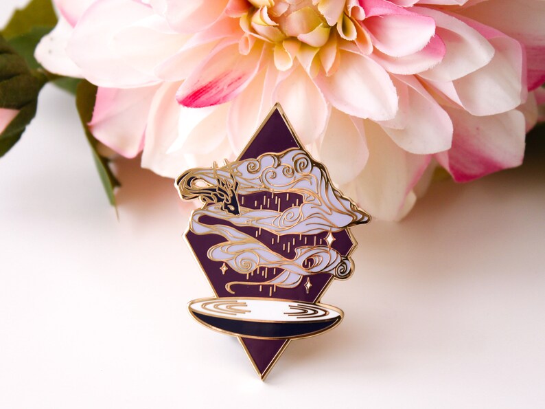 Pu&ograve; includere: Uno spillo in smalto dorato con uno sfondo a forma di diamante viola. Lo spillo presenta un design bianco e oro di nuvole vorticose e gocce di pioggia sopra un piatto circolare bianco e nero.