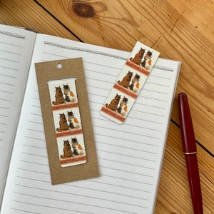 May include: Two cat-themed bookmarks and a matching bookmark card are displayed on a lined notebook. The bookmarks feature illustrations of two cats sitting together. The cats are brown, white, and orange. A red pen rests on the notebook.