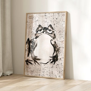 May include: A framed art print featuring a black and white ink illustration of a frog. The frog has a contemplative expression. The background is filled with Asian calligraphy. The frame is made of light-colored wood.