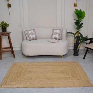 Puede incluir: Una alfombra rectangular de yute marr&oacute;n claro con un borde tejido. La alfombra se coloca delante de un sof&aacute; blanco con dos almohadas y un libro encima. Una planta en maceta est&aacute; en el fondo.