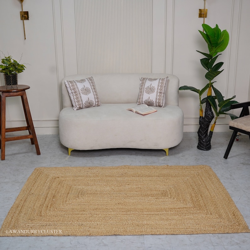 Puede incluir: Una alfombra rectangular de yute marr&oacute;n claro con un borde tejido. La alfombra se coloca delante de un sof&aacute; blanco con dos almohadas y un libro encima. Una planta en maceta est&aacute; en el fondo.
