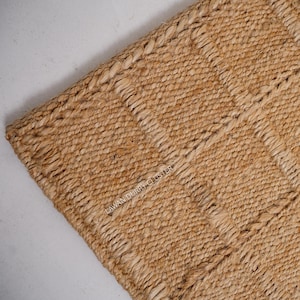 May include: Close-up of a natural jute rug with a woven texture. The rug has a light brown color with darker brown stripes and a braided edge. The text "LAWAN DURRY CLUSTER" is visible on the rug.