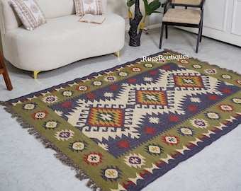 May include: A rectangular area rug featuring a dark blue center with a repeating geometric pattern in cream, olive green, red, and purple. The border is olive green with small geometric motifs and fringed edges. The text RugsBoutiques is visible.