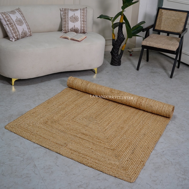 Puede incluir: Una alfombra de fibra natural con forma cuadrada y un borde tejido. La alfombra est&aacute; enrollada en el fondo y la imagen muestra la alfombra extendida sobre un suelo gris claro.