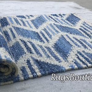May include: A rolled blue and white patterned rug. The rug has a geometric design with blue diamond shapes and white accents. The rug is made of a textured material and is partially rolled up, showing its pattern and texture.