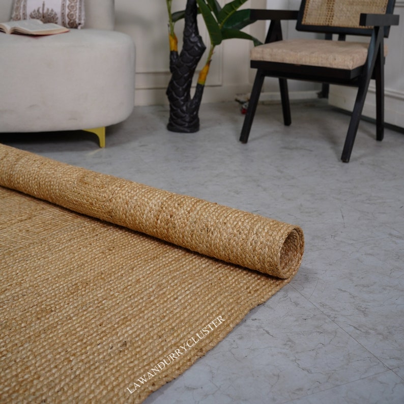 Puede incluir: Una alfombra de fibra natural enrollada de color marr&oacute;n claro. La alfombra est&aacute; hecha de yute tejido y tiene una superficie texturizada. La alfombra est&aacute; parcialmente desenrollada sobre un suelo gris claro.