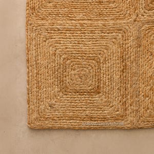 May include: Close-up of a natural jute rug with a geometric square pattern. The rug is a warm tan colour and has a woven texture. The design features concentric squares, creating a textured surface. Suitable for home decor.