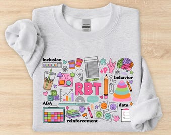 May include: A light gray crewneck sweatshirt featuring a colorful graphic print with the text RBT, inclusion, behavior, ABA, data, and reinforcement. The design includes illustrations of a brain, a stack of rings, a clipboard, and various icons.