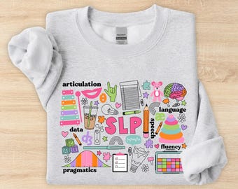 May include: A light gray crewneck sweatshirt featuring a colorful graphic print with the text SLP, articulation, language, data, pragmatics, and speech. The design includes illustrations of a xylophone, brain, pencil, clipboard, and various shapes.