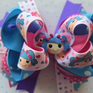 May include: A large, colorful hair bow featuring a cartoon doll design. The bow is made of layered ribbons in shades of blue, pink, purple, and white, with polka dots and heart patterns. A doll figure is centered on the bow.